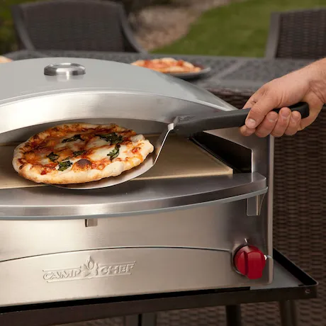 Eine Pizza, die mit einer Pizzaschaufel in einen schlanken, aus Edelstahl gefertigten Outdoor-Pizzaofen gelegt wird. Die Pizza ist mit Käse und Basilikum belegt, und der Ofen steht auf einer Terrasse.